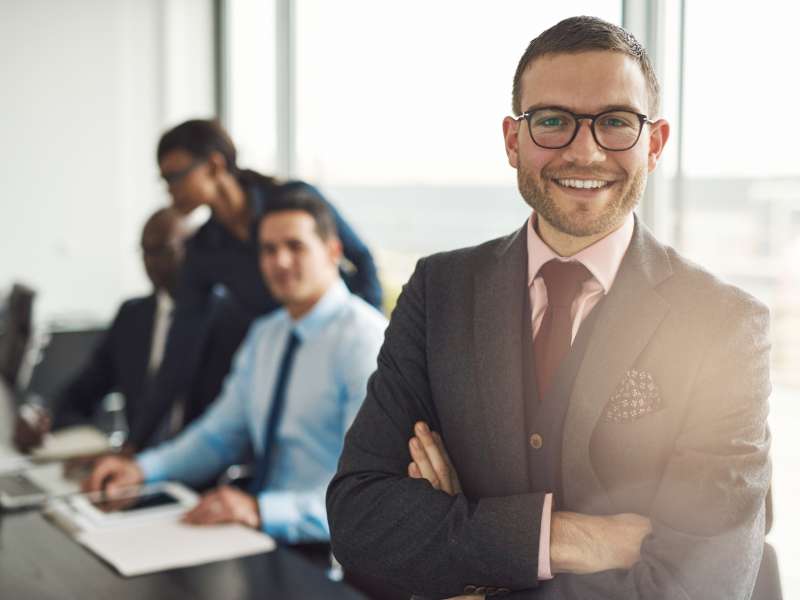 Confident smiling business executive with folded arms near conference table with three co-workers discussing something in large bright office room